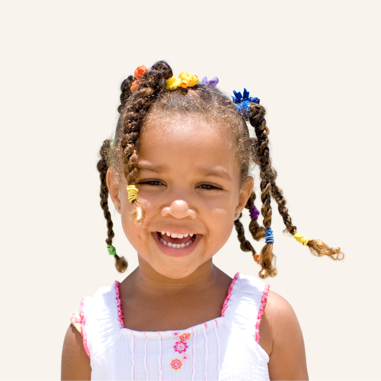 Smiling young girl with braided hair decorated with colorful hair ties, wearing a white dress with pink embroidery, against a plain light background.