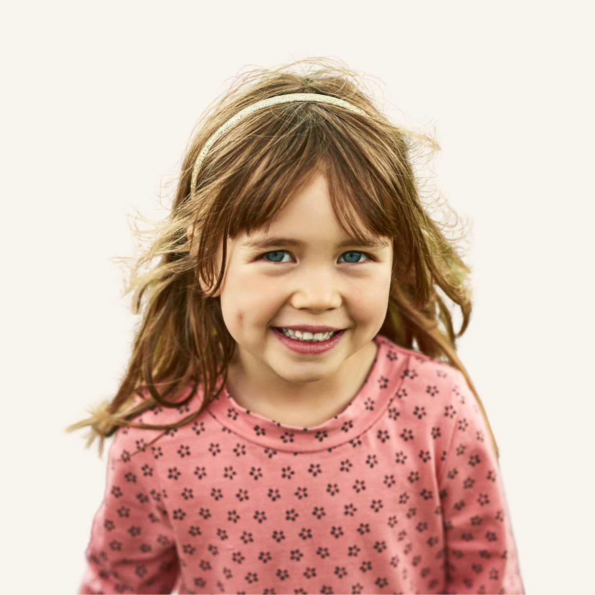 Smiling young girl with long brown hair and blue eyes, wearing a pink patterned shirt and a headband, standing against a plain light background.