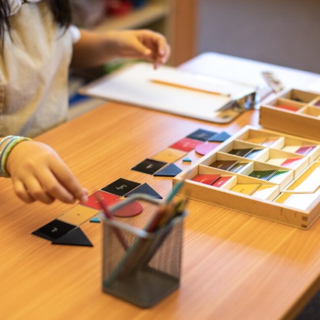 A child sits at a desk arranging geometric shapes and using colored tiles, with a pencil holder and notepad nearby.