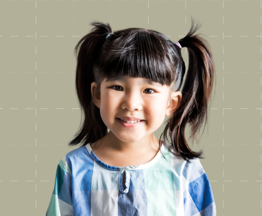 Young girl with dark pigtails and bangs, wearing a blue and white checkered shirt, smiling in front of a plain, light-colored background.