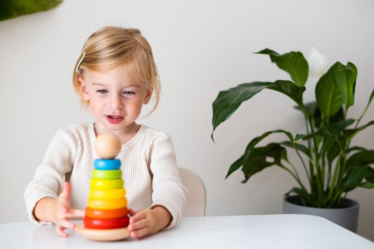 A young child sitting at a table stacking colorful wooden rings, with a green potted plant in the background.