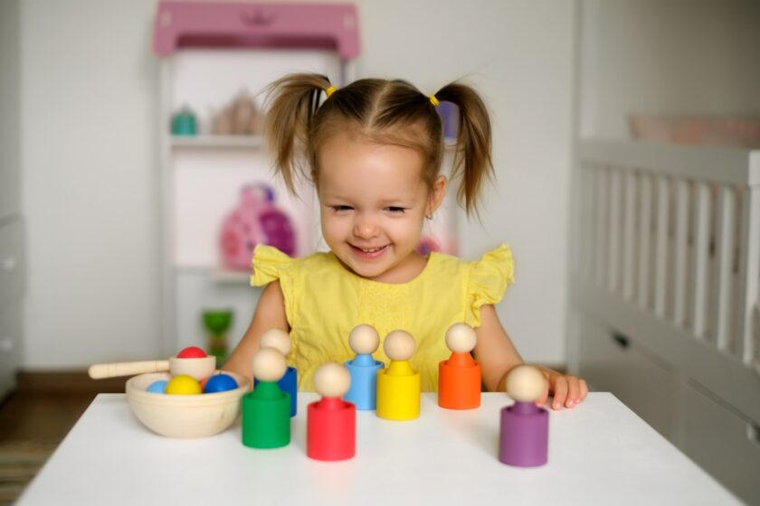 A young girl in a yellow dress smiles while playing with colorful wooden figures on a white table in a bright room.
