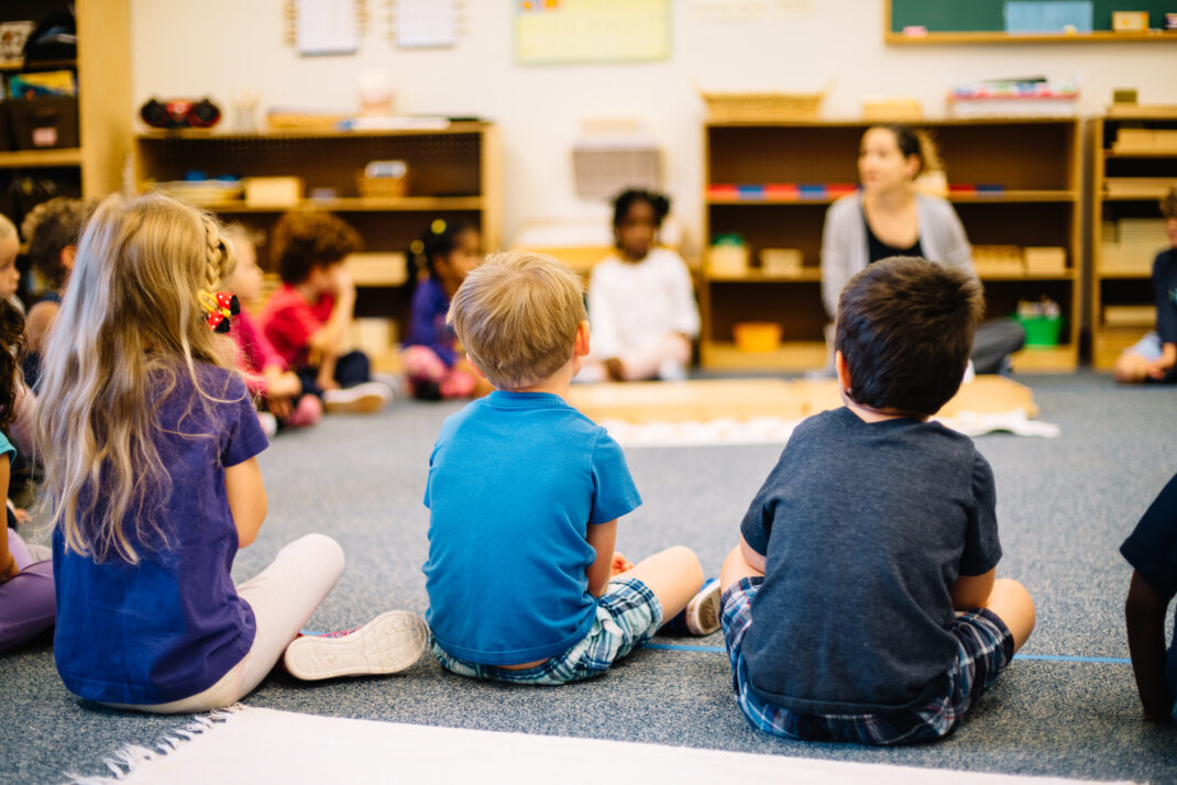 Young children sit in a circle on the classroom floor, facing a teacher who is leading an activity. Shelves with educational materials are visible in the background.