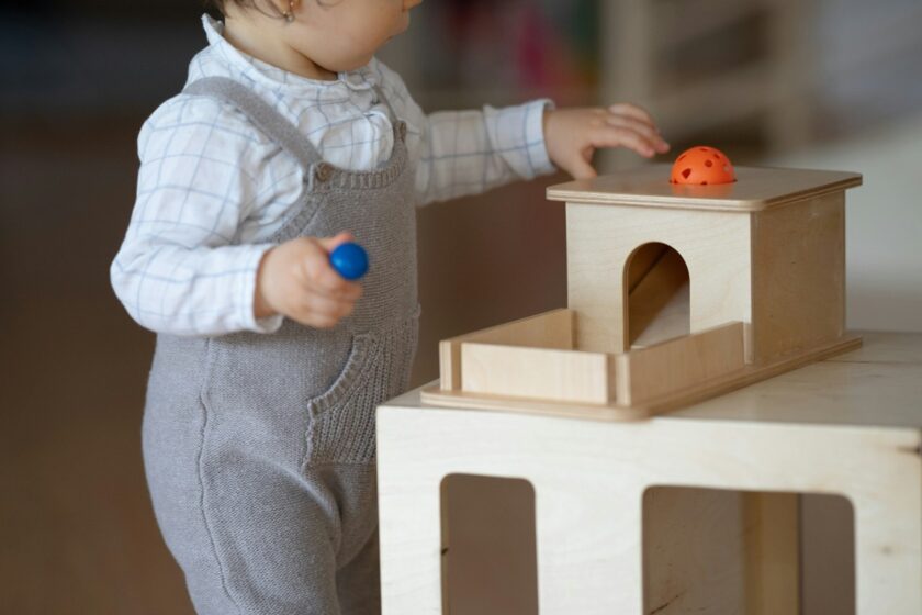 A toddler in gray overalls plays with a wooden toy, placing a ball on top and holding another ball in the other hand.