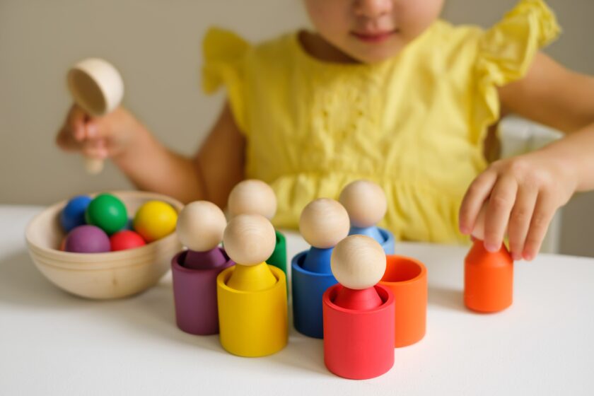 A child in a yellow dress plays with colorful wooden peg dolls and matching cups, sorting them by color on a white table.