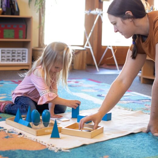 An adult and a young girl sit on a colorful map rug, arranging blue geometric shapes on a white mat in a classroom setting.