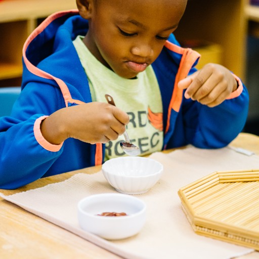 A young child sitting at a table uses a spoon to scoop from a small white bowl, with another bowl and a tray placed nearby.
