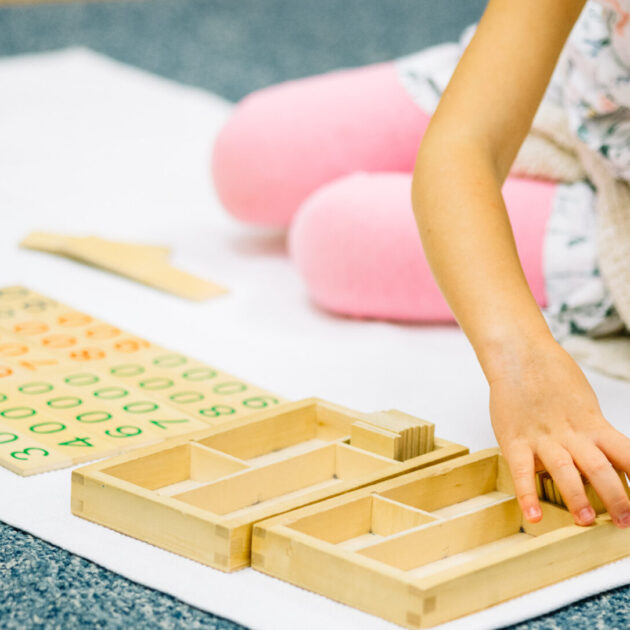 A child sits on the floor using wooden Montessori math materials with numbers and counting trays placed on a white mat.