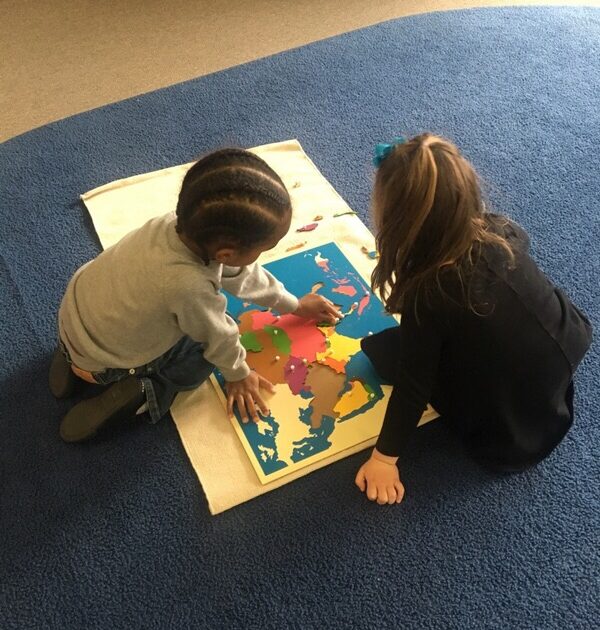 Two young children sit on a blue carpet, working together on a colorful world map puzzle placed on a white mat.
