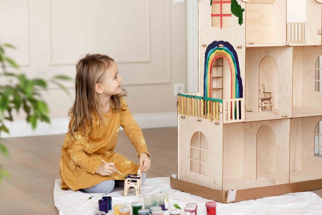 A young girl in a yellow dress paints a small wooden chair and a rainbow on a large wooden dollhouse, with paint jars and a brush on the floor.