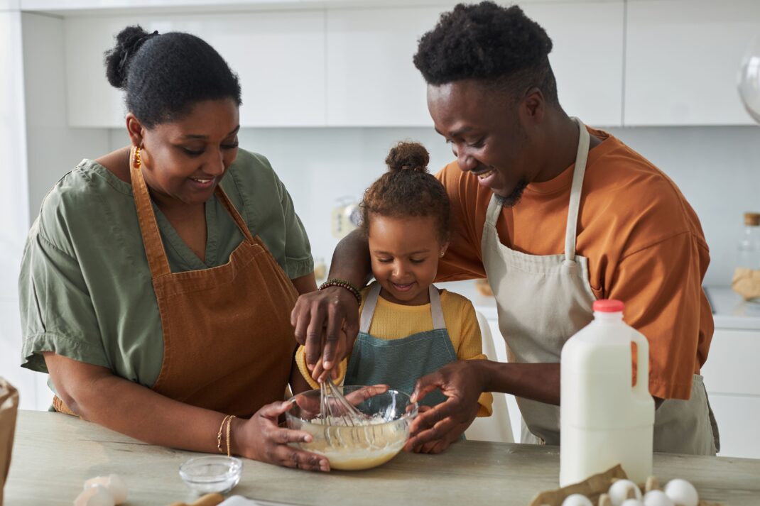 Three people, two adults and a child, are standing in a kitchen mixing ingredients in a bowl, wearing aprons and smiling. A bottle of milk and eggs are on the counter.