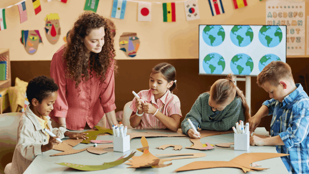 A teacher supervises four children working on a craft project at a classroom table decorated with international flags and cut-out materials.