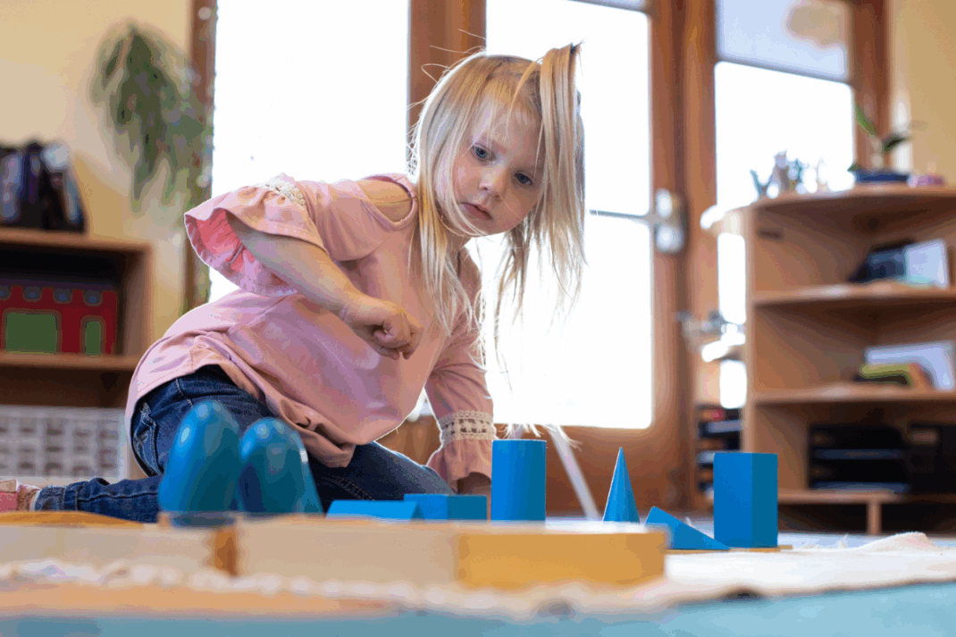 Young girl in a pink shirt sits on the floor indoors, engaging with blue geometric shapes on a mat in a brightly lit room.