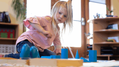 Young girl in a pink shirt sits on the floor indoors, engaging with blue geometric shapes on a mat in a brightly lit room.