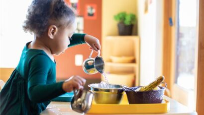 A young child pours water from a metal cup into a bowl at a table, with kitchenware and a few bananas nearby.