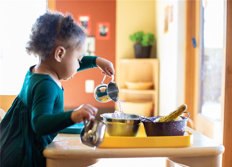A young child pours water from a metal cup into a bowl at a table, with kitchenware and a few bananas nearby.